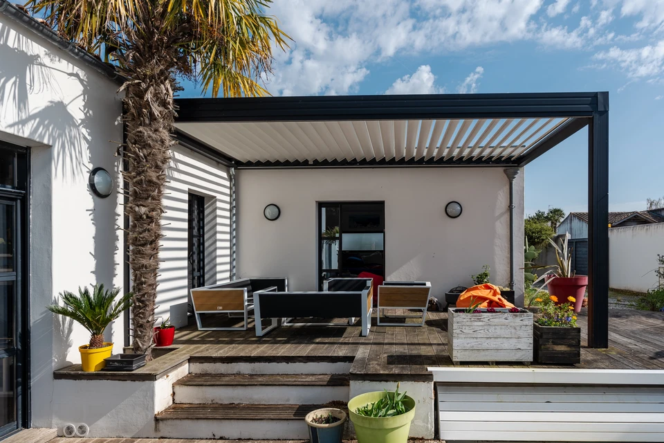 Terrasse couverte moderne avec pergola bioclimatique à lames orientables noires, mobilier d'extérieur gris contemporain, vue sur villa en pierre naturelle et végétation méditerranéenne