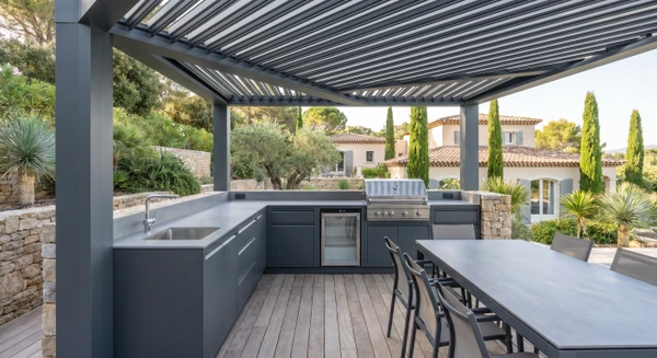 Terrasse extérieure moderne avec cuisine d'été entièrement équipée et intégrée en structure aluminium gris anthracite : comptoir de cuisine avec plan de travail, évier encastré, espace de rangement, mini-réfrigérateur et barbecue intégré. La cuisine est couverte par une pergola bioclimatique ou une structure d'ombrage. Mobilier de relaxation et espace repas visible à proximité. Environnement résidentiel haut de gamme, lumière naturelle du jour, terrasse dallée ou en bois, démontrant l'intégration harmonieuse et la qualité de fabrication.